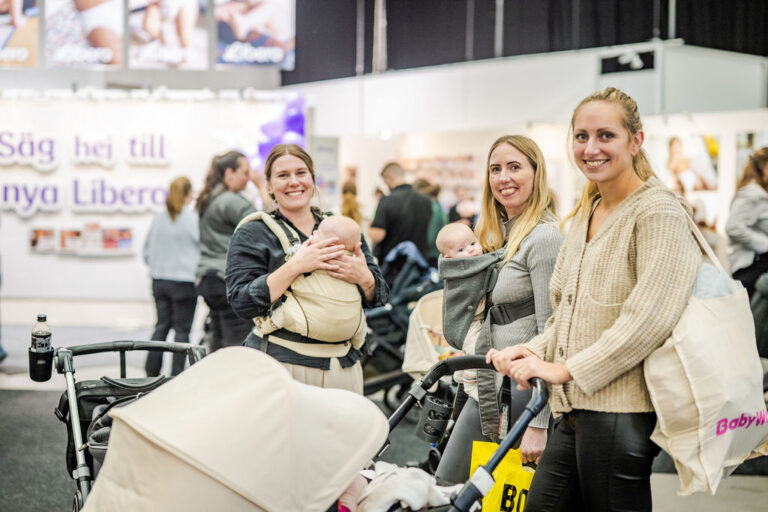 En grupp glada föräldrar som bär sina bebisar i bärselar och står bredvid barnvagnar på Underbara barn-mässan.