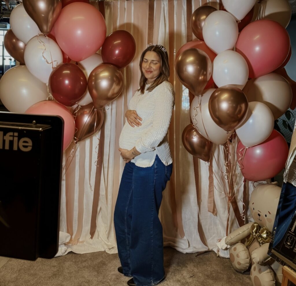 En gravid kvinna som poserar framför en dekorativ fotovägg på Mom shower-mässa.